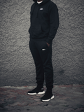 A person wearing black joggers and a hooded sweatshirt, standing against a gray background.