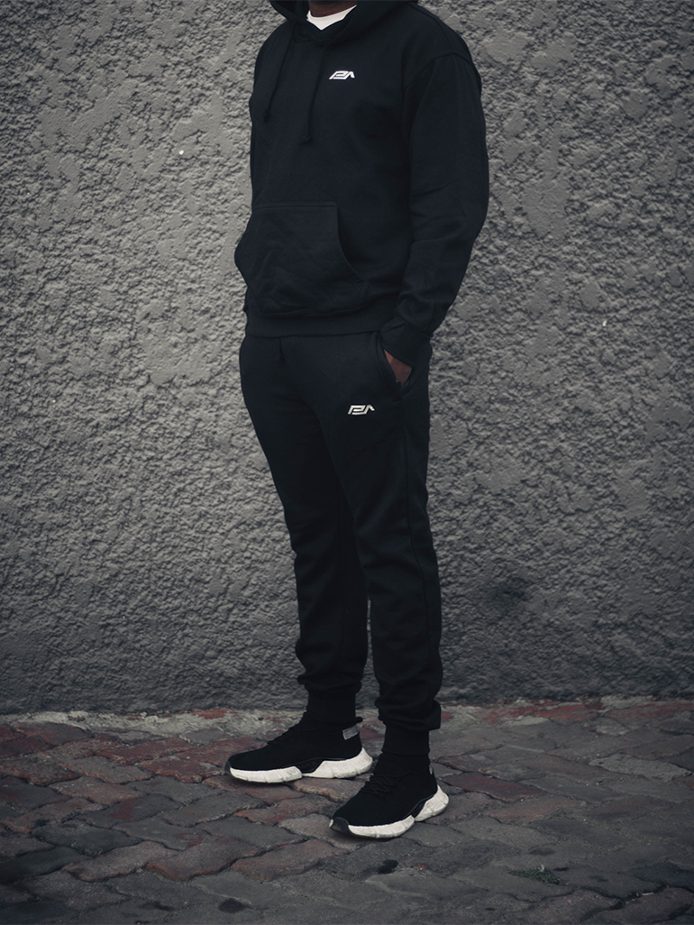A person wearing black joggers and a hooded sweatshirt, standing against a gray background.